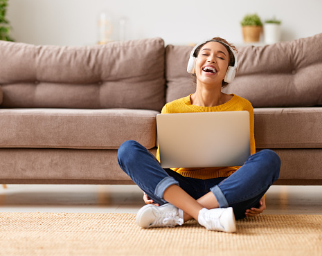 Young happy afro american woman, student or office worker, wearing headphones sitting on floor and listening music, enjoying favourite audio track while working or styding online on laptop at home