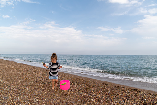 Photo of 2,5 yeas old girl playing on beach sand. Sky is partly cloudy. She is barefoot. A pink colored bucket is seen next to her. Shot in outdoor day light with a full frame mirrorless camera.