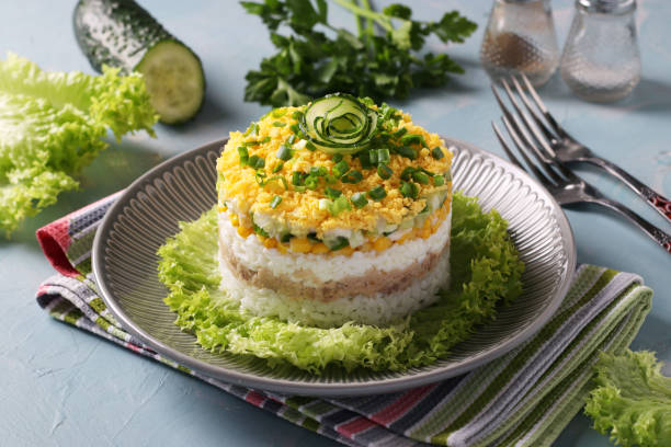 istock Festive puff salad with cod liver, rice, eggs, corn and cucumber on a plate on light blue background. Close up 1315053883