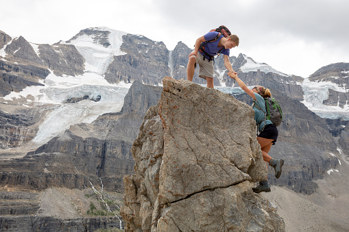 He offers her a helping hand up cliff, Canadian Rockies in the distance