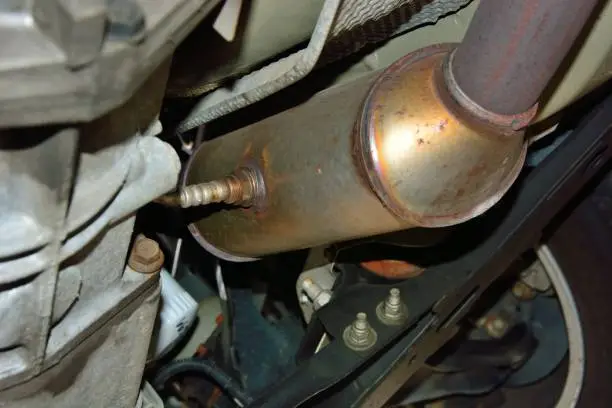 Selective focus on a stainless steel catalytic converter with an oxygen sensor or O2 sensor installed on an exhaust system under a vehicle. Catalytic converters have become a target for theft.