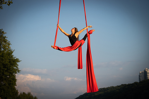 Beautiful and flexible female circus artist dancing with aerial silk on a sky background.