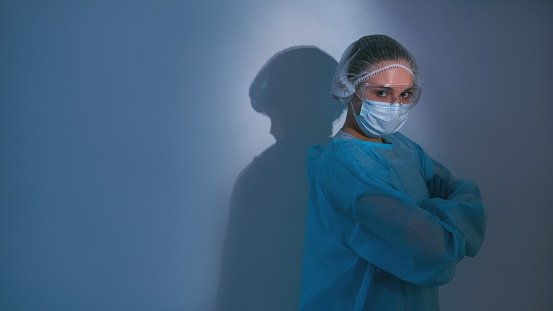Covid-19 protection. Pandemic banner. Healthcare worker. Quarantine hygiene. Portrait of confident brave female doctor hero in ppe face mask goggles with crossed arms on blue copy space background.