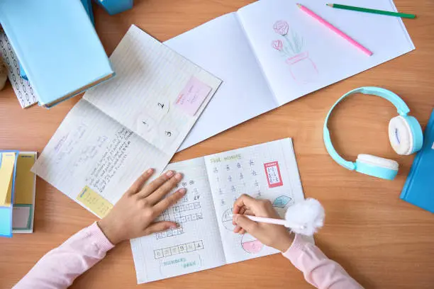 Photo of Table top closeup view of kid student hands writing doing math homework exercises in notebook with book on table. Learning classes for elementary school pupils. Study at home classroom concept.