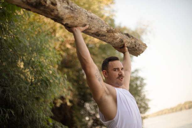 Bodybuilder on the beach picks up a tree and exercises.