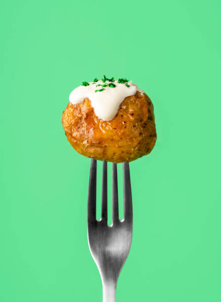 Roasted baby potatoes stuck on a fork, isolated on a green background. Single roasted potato with sour cream sauce and herbs