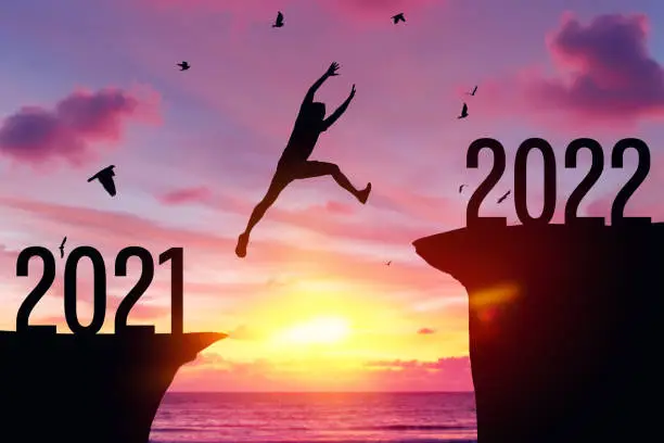 Photo of Silhouette man jumping between cliff with number 2021 to 2022 and birds flying at tropical sunset beach.