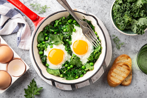 Healthy breakfast. fried eggs with kale, spinach and green peas in red skillet , top view