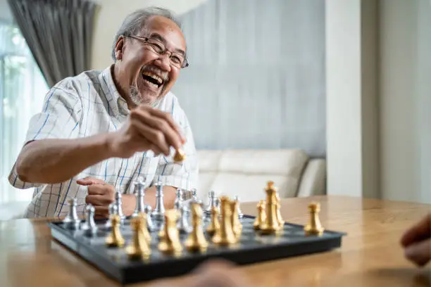 Photo of Portrait of Asian Senior Elderly male spend leisure time, stay home after retirement. Happy smiling Old man enjoy activity in house play chess game with friend. Hospital Healthcare and medical concept