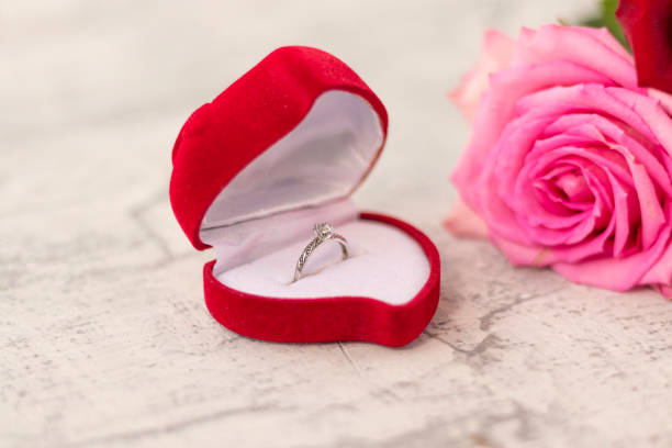 wedding ring with diamond in a red heart-shaped box and a bouquet of roses on a white textured background. Concept for engagement, gift, anniversary, love, wedding or valentine's day stock photo