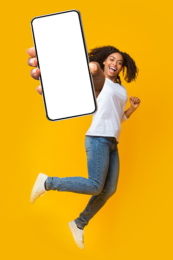 Cheerful African American woman jumping in air, having fun, showing smartphone with empty white screen over orange studio background, mockup for mobile app or website. Collage