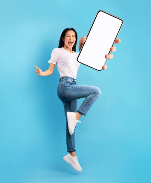 istock Advertisement for mobile app. Excited lady jumping over blue background, showing modern smartphone with empty screen 1315129141