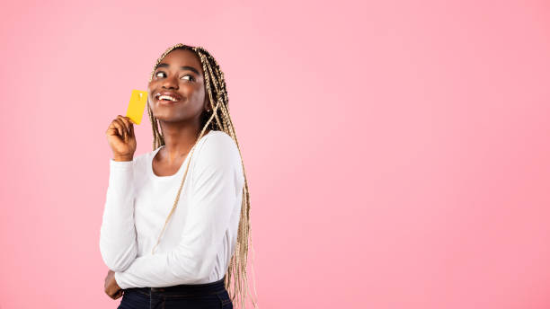 Black woman holding credit card and dreaming What To Buy. Portrait of black woman holding credit card and looking back at copy space, thinking about purchase, dreaming about gifts, standing isolated on pink studio background, banner, panorama thinking of you card stock pictures, royalty-free photos & images