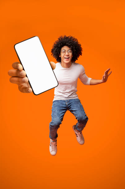 istock Mobile app advertisement. Excited teen black guy showing smartphone with empty screen, jumping on orange background 1315129527