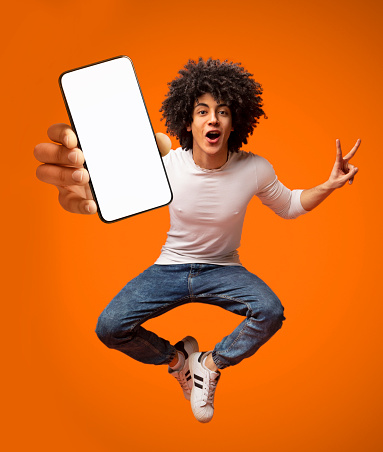 Portraif of excited African American teen guy jumping with smartphone, demonstrating empty screen on orange background, copy space for your mobile advertisement, mockup image