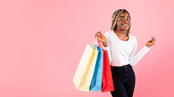 Shopaholic Concept. Portrait of excited young black woman with afro braids holding colorful shopping bags from mall, looking back at free copy space, pink studio background, banner, panorama