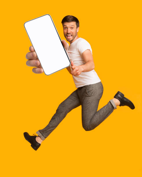 istock Excited man jumping, pointing finger at camera, showing smartphone with blank screen over orange background, mockup 1315136164