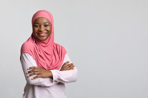 Smiling young black lady in pink hijab posing on grey studio background, panorama with copy space. Cheerful african american millennial woman standing next to empty space for advertisement