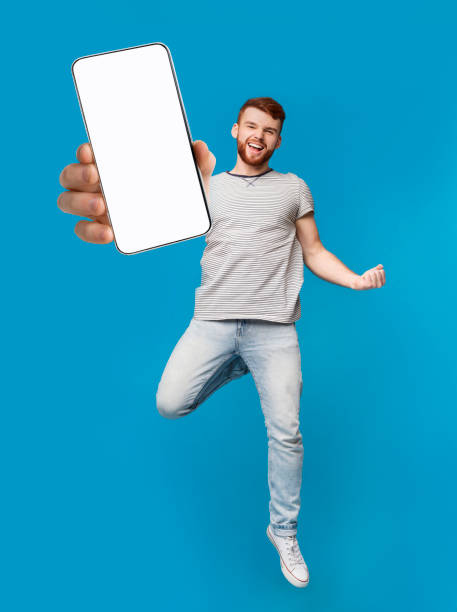 istock Excited redhead man jumping with smartphone, showing YES gesture on blue studio background, mockup for mobile app 1315136897