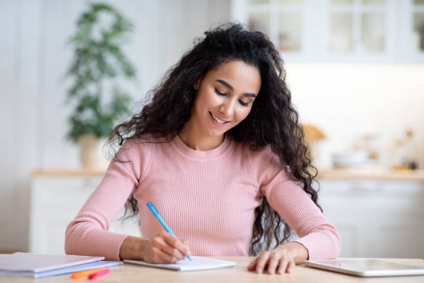 istock Young Smiling Woman Writing Notes In Notepad, Sitting At Table In Kitchen 1315137581