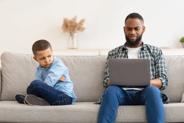 Father's Indifference. Black Dad Ignoring Offended Son Using Laptop Sitting On Couch Indoors. Reluctant Daddy Neglecting His Unhappy Kid. Bad Relationship With Father Concept