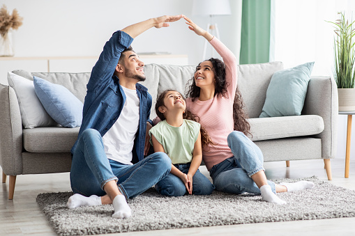 Family Insurance Concept. Young Arab Parents Making Roof Symbol Of Hands Above Their Little Daughter While Sitting On Floor At Home, Middle Eastern Mom, Dad And Child Enjoying New House, Free Space