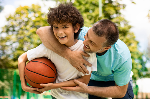 Enjoying Game. Portrait of cheerful father and son playing basketball together, smiling positive boy holding ball and looking at camera, excited man embracing him from behind and trying to take ball