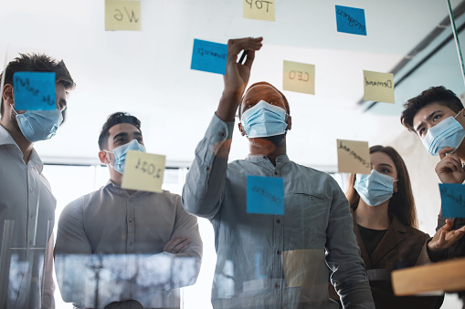 Workshop And Brainstorming Concept. Diverse group of pensive multicultural business people in masks having meeting, using sticky paper post-it notes on glass wall or window to share ideas and thinking