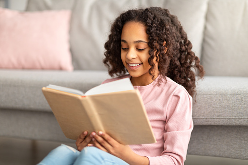 Happy African girl reading paper book with pleasure, sitting on the floor near sofa at home
