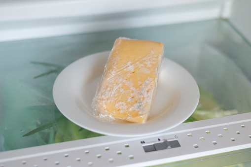 Moldy cheese wrapped in cling film in the refrigerator. Incorrect storage, spoiled product, expired concept.