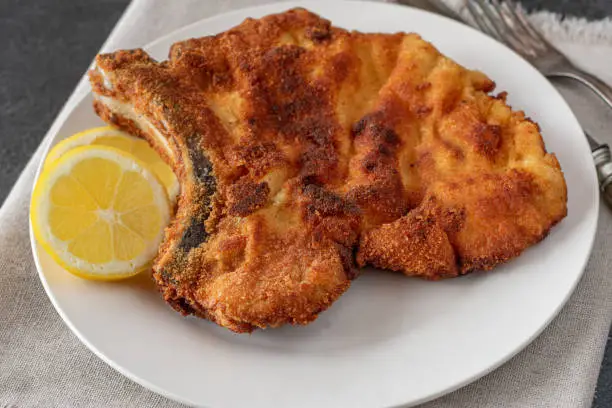 Delicious homemade pan fried cutlet or pork chop with breading served with sliced lemon on a white plate on kitchen table background from above