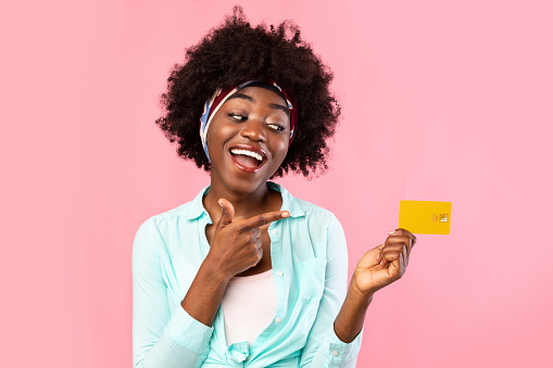 Cheerful Black Woman Pointing Finger At Credit Card Recommending Banking Service Posing In Studio Over Pink Background. Money And Finances, Payment And Shopping Concept