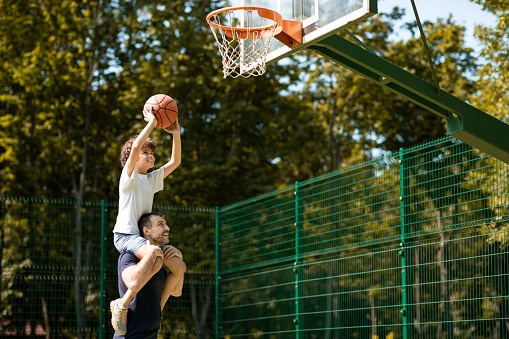 Happy Loving Family. Portrait of young curly boy sitting on father's shoulders, playing basketball, throwing ball to basket, man standing on court outdoors and helping his son, selective focus