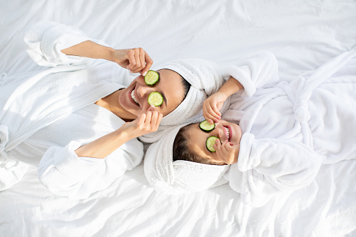 Top view of playful mother and daughter in bathrobes and white towels on heads laying on bed, holding cucumber circles, making face care procedures at home. Motherhood, parenthood concept