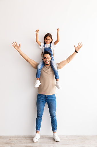 Happy Family. Full Body Length Vertical Portrait Of Cheerful Man Riding Excited Daughter On Shoulders, Looking And Posing At Camera, Having Fun, Raising Hands Up Standing Isolated Over Wall At Home