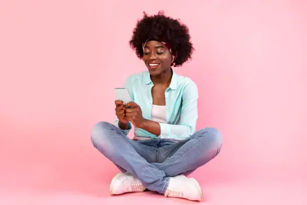 Happy African Woman Using Phone Texting And Browsing Internet Sitting On Floor Over Pink Background. Bushy Black Lady Networking In Social Media Using Smartphone App. Mobile Application For Cellphone