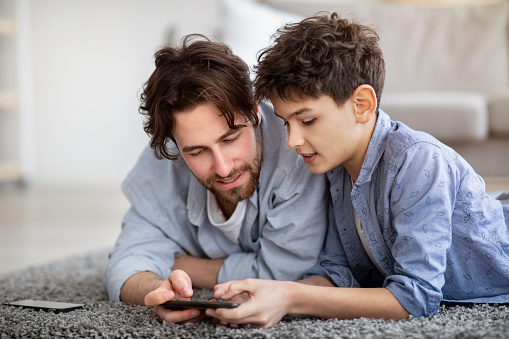Father and son using smartphone, watching photos or boy helping daddy with game, spending time at home while lying on floor carpet. Modern technologies and leisure activities concept