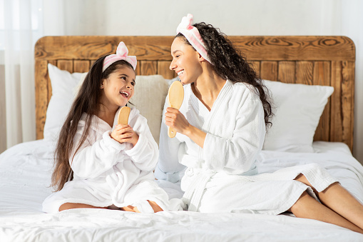 Funny fair-haired mother and daughter with wooden hair brushes singing at home, wearing white bathrobes and hair bands, sitting on bed, happy family having fun together, copy space