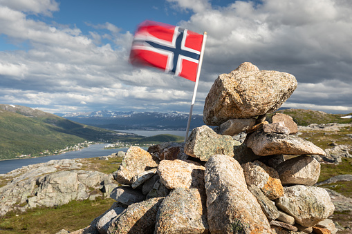Norwegian flag in the wind at mountains top. Rocks, fjord, city of Tromso and snowy mountains in background