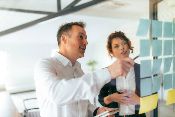 istock Discussing ideas. Two business people talking while looking at post it notes on a glass wall. 1315151377