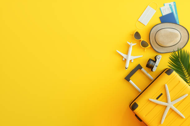 Reisepass, Flugtickets, gelber Koffer, Sonnenhut und schutzbeschützende Gesichtsmaske auf gelbem Hintergrund