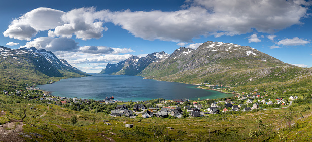 Summer fjord panorama with village at end of fjord. Mountains covered with snow and beautiful clouds over sea