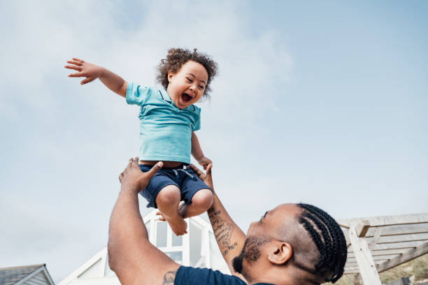 Young boy with Down Syndrome expressing joy in mid-air Over the shoulder view of 35 year old Black father lifting ecstatic 4 year old son high into the air as they enjoy outdoor playtime together. special needs child pictures stock pictures, royalty-free photos & images