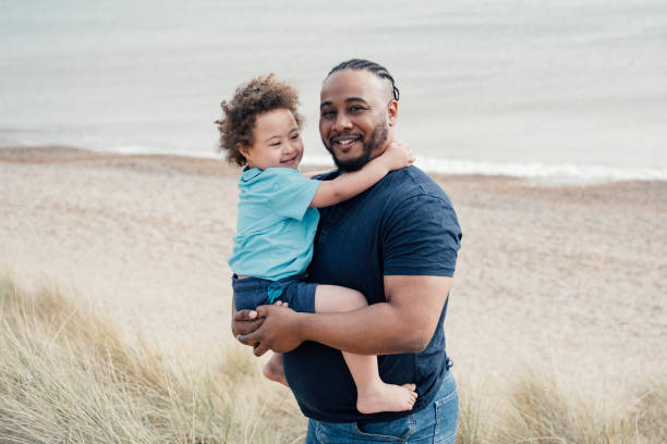 Portrait of British father and son vacationing at the beach Waist-up view of cheerful Black man in mid 30s holding smiling 4 year old son with Down Syndrome on dune in Camber Sands with Rye Bay in background. down syndrome photos stock pictures, royalty-free photos & images