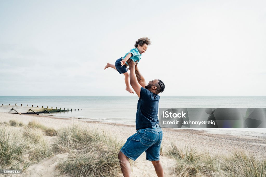 Father and son enjoying vacation playtime at Camber Sands Side view of mid adult British man standing on dune lifting excited 4 year old boy with Down Syndrome high into the air with Rye Bay in background. Beach Stock Photo