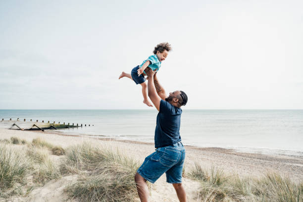 Father and son enjoying vacation playtime at Camber Sands Side view of mid adult British man standing on dune lifting excited 4 year old boy with Down Syndrome high into the air with Rye Bay in background. disability photos stock pictures, royalty-free photos & images