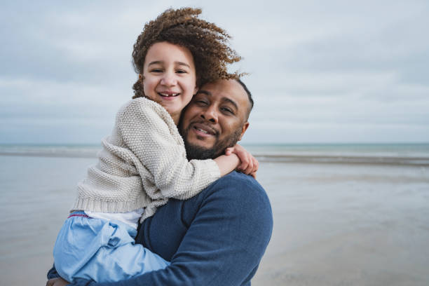 Portrait of British father and daughter at Camber Sands Close-up of happy and windblown mid 30s Black man holding 6 year old daughter in his arms as they explore the beach at low tide with Rye Bay in background. gap toothed photos stock pictures, royalty-free photos & images