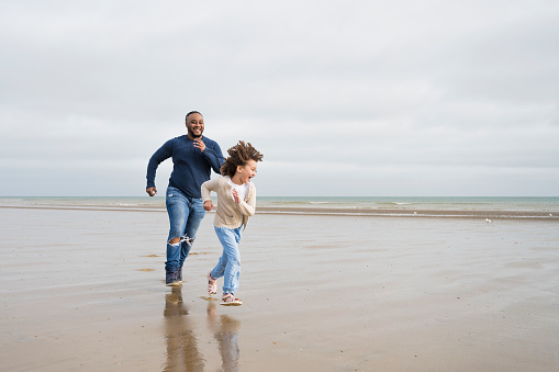 Front view of Black man in mid 30s and mixed race 6 year old daughter running and laughing as they chase each other near water’s edge at low tide.