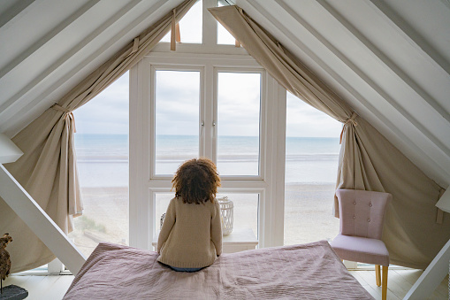 Rear view of 6 year old girl with curly brown hair sitting on end of bed looking at Rye Bay on southeast coast of England from second story bedroom.