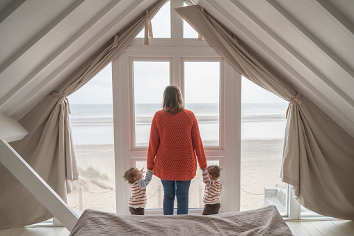 Rear view of mid adult woman holding hands with 23 month-old daughters and standing at second story window looking at view of sand and sea.